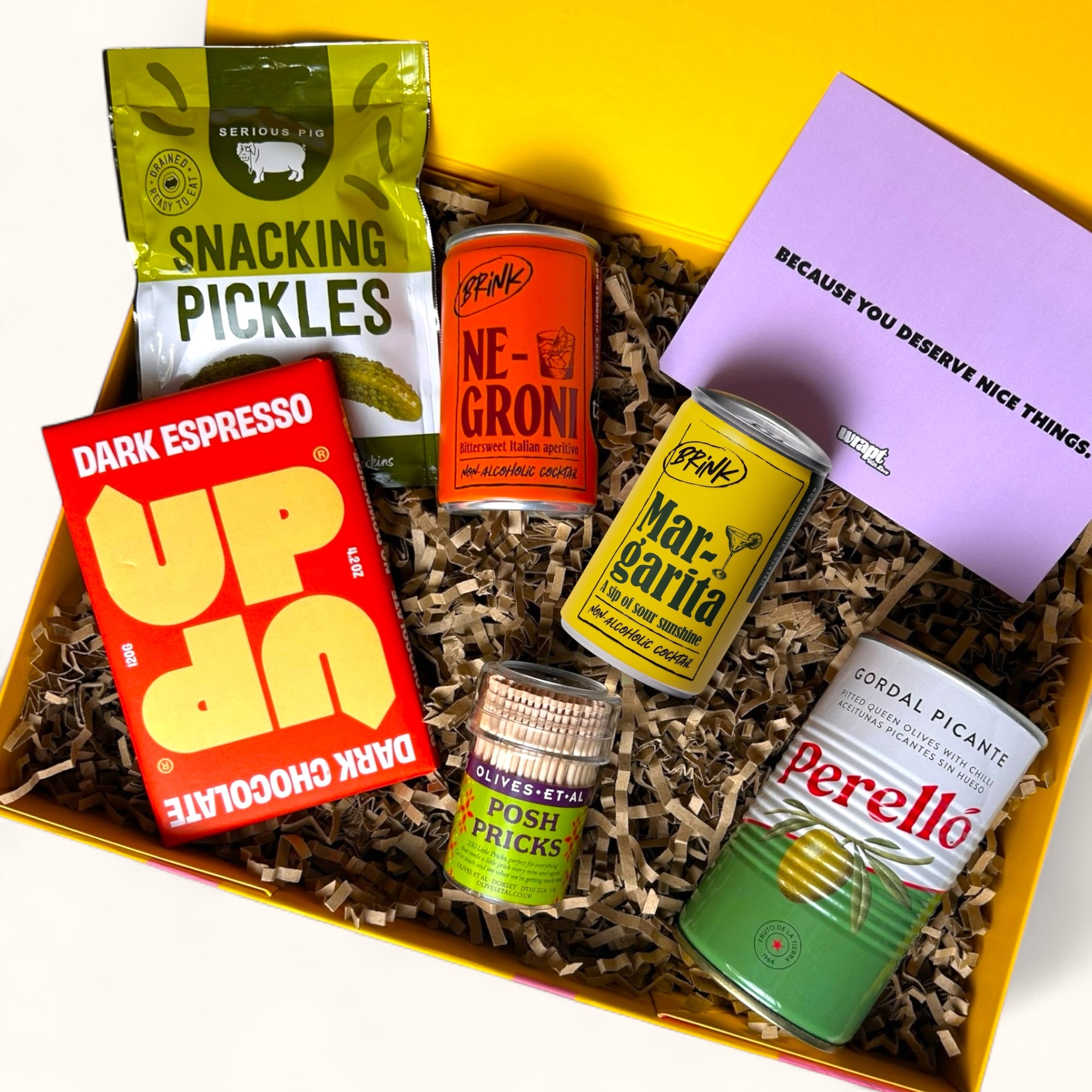 Bright yellow gift box filled with alcohol free cockktails, snacks and cocktail sticks.