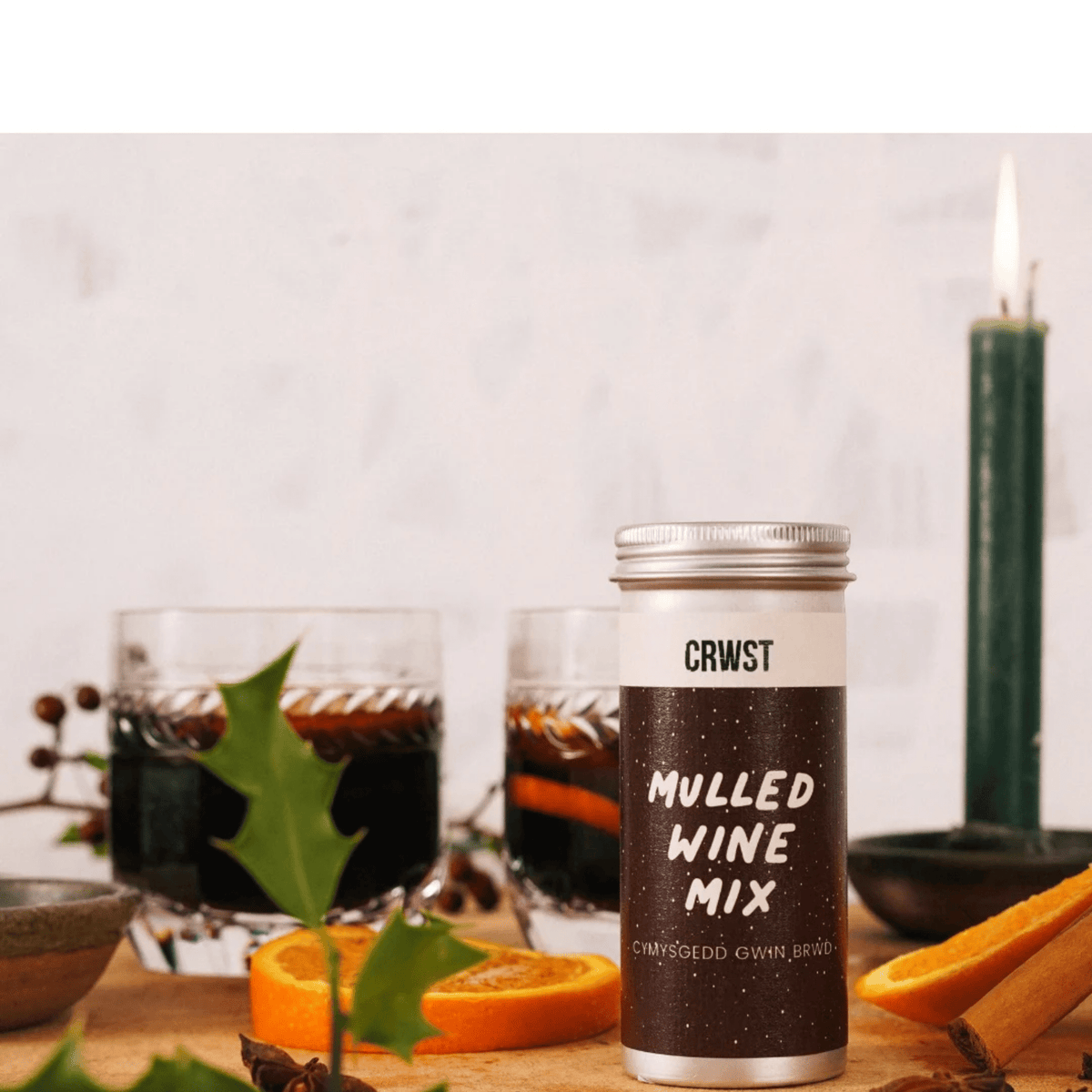 A bottle of CRWIST Charred Own Brand Mulled Wine Mix stands on a wooden surface, styled with two glasses of dark mulled wine, orange slices, cinnamon sticks, a green candle, and a blurred background.