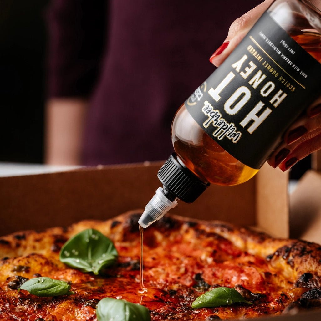 Person drizzling honey from a bottle onto a pizza with basil leaves.